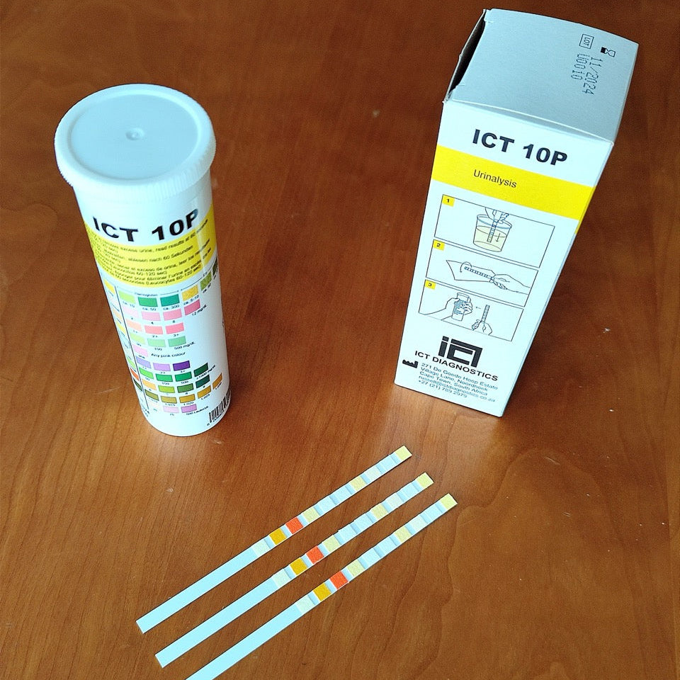 ICT 10P urine test strips and their packaging on a wooden table, including a white plastic vial with a color chart, a white and yellow retail box, and three test strips.