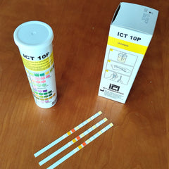 ICT 10P urine test strips and their packaging on a wooden table, including a white plastic vial with a color chart, a white and yellow retail box, and three test strips.